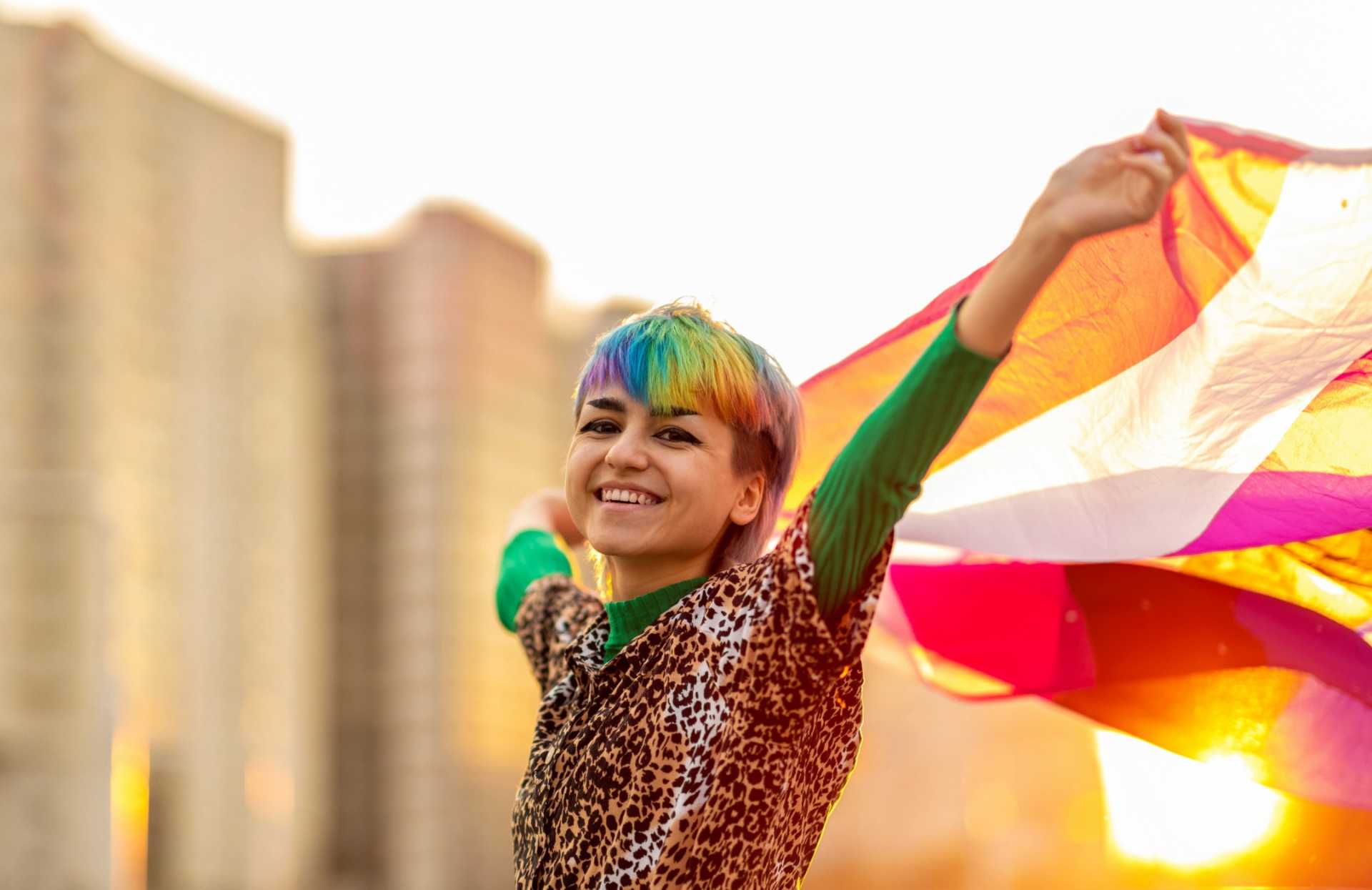 A non-binary person with rainbow coloured hair holding a rainbow flag