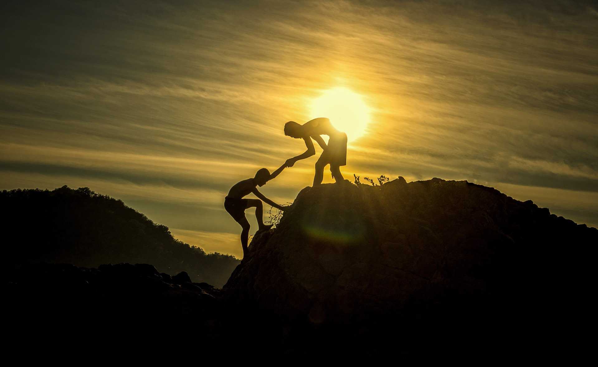 Two people helping each other up a mountain with sunset in the background