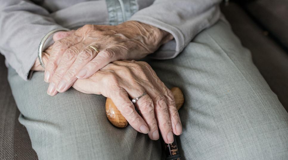 Older person with hands crossed on lap and walking aid in hand